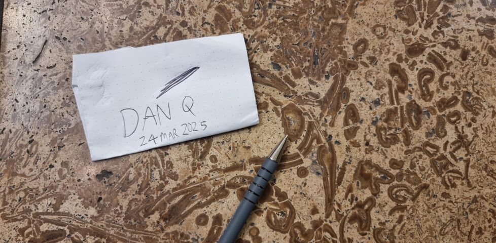 "Dan Q" and today's date written on a small piece of paper, alongside a pen, which points to a bivalve fossil in a limestone floor.