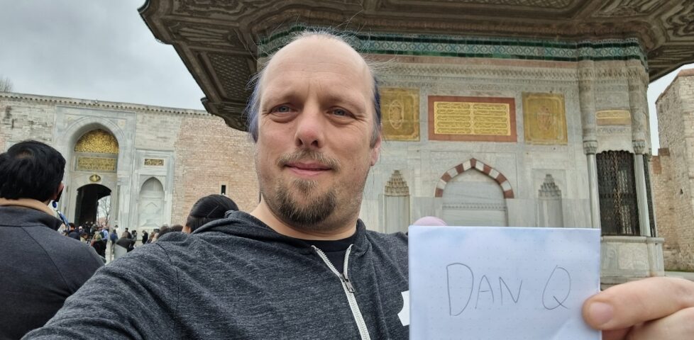 Dan holds up a sign showing his name in front of a gilded fountain decorated in Arabic script.