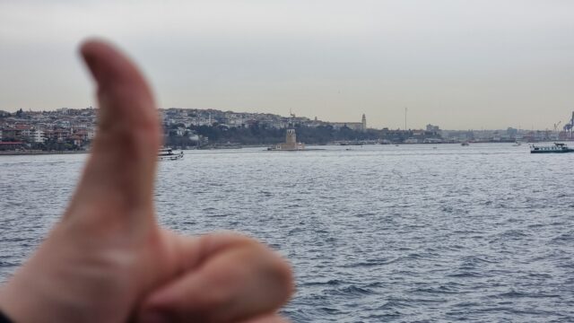 A "thumbs up" sign close to the lens, looking over the water towards a tower, close to shore.