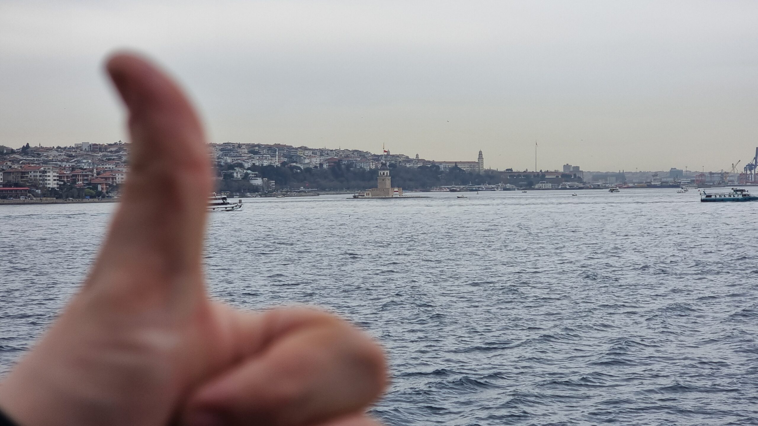 Full image of A "thumbs up" sign close to the lens, looking over the water towards a tower, close to shore.