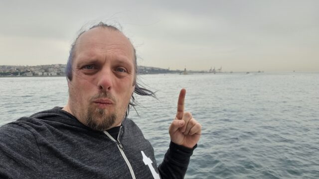 Dan stands and takes a selfie while pointing to the distant tower.