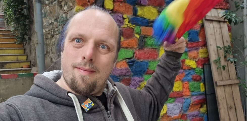 Dan waves a rainbow bandana in front of a wall painted in multitudinous colours, on a staircase with each step painted a different colour.