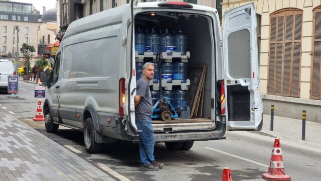 A grey-haired man wearing a t-shirt and blue jeans leans casually against the open doors of a white van that's visibly filled floor-to-ceiling with 19-litre 'water cooler style' water bottles, plus a pallet lifter. The van is parked between red-and-white striped cones on an Istanbul street.