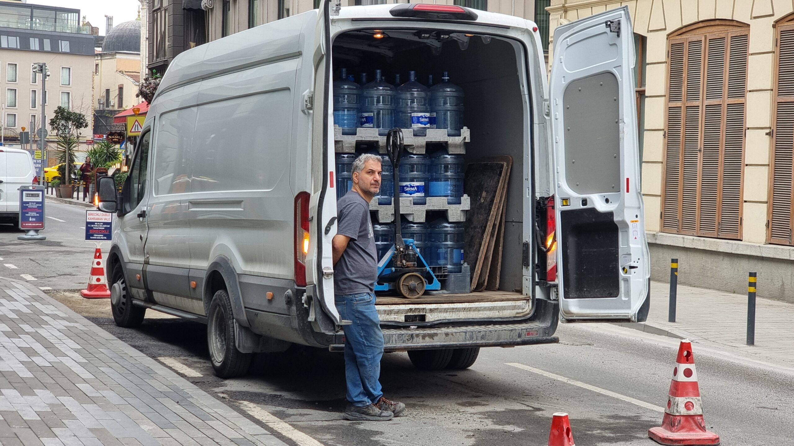 Full image of A grey-haired man wearing a t-shirt and blue jeans leans casually against the open doors of a white van that's visibly filled floor-to-ceiling with 19-litre 'water cooler style' water bottles, plus a pallet lifter. The van is parked between red-and-white striped cones on an Istanbul street.