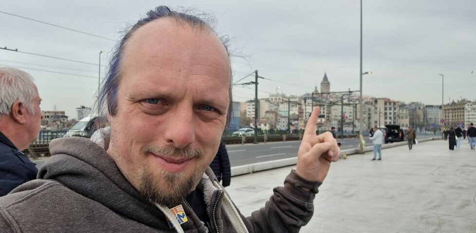 Dan stands on a busy, wide foot/road bridge, pointing at the top floor of a building overlooking the river that it crosses.