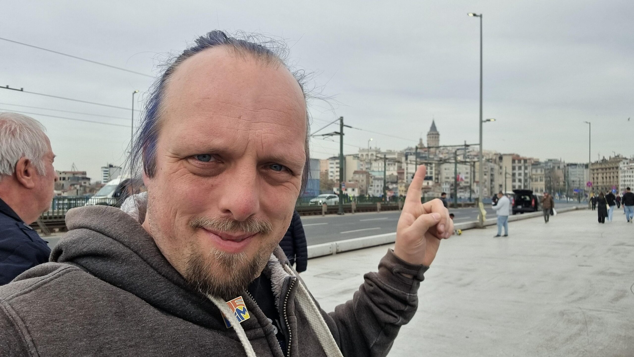Full image of Dan stands on a busy, wide foot/road bridge, pointing at the top floor of a building overlooking the river that it crosses.