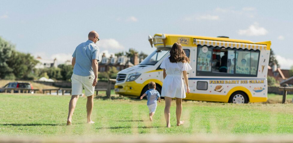 On a verdant manicured lawn under a summer sky, two parents walk barefoot after their young child, who is running towards a traditionally-coloured yellow-and-white British ice cream van with visible branding reading 'fresh dairy ice cream' and '99 flake'.