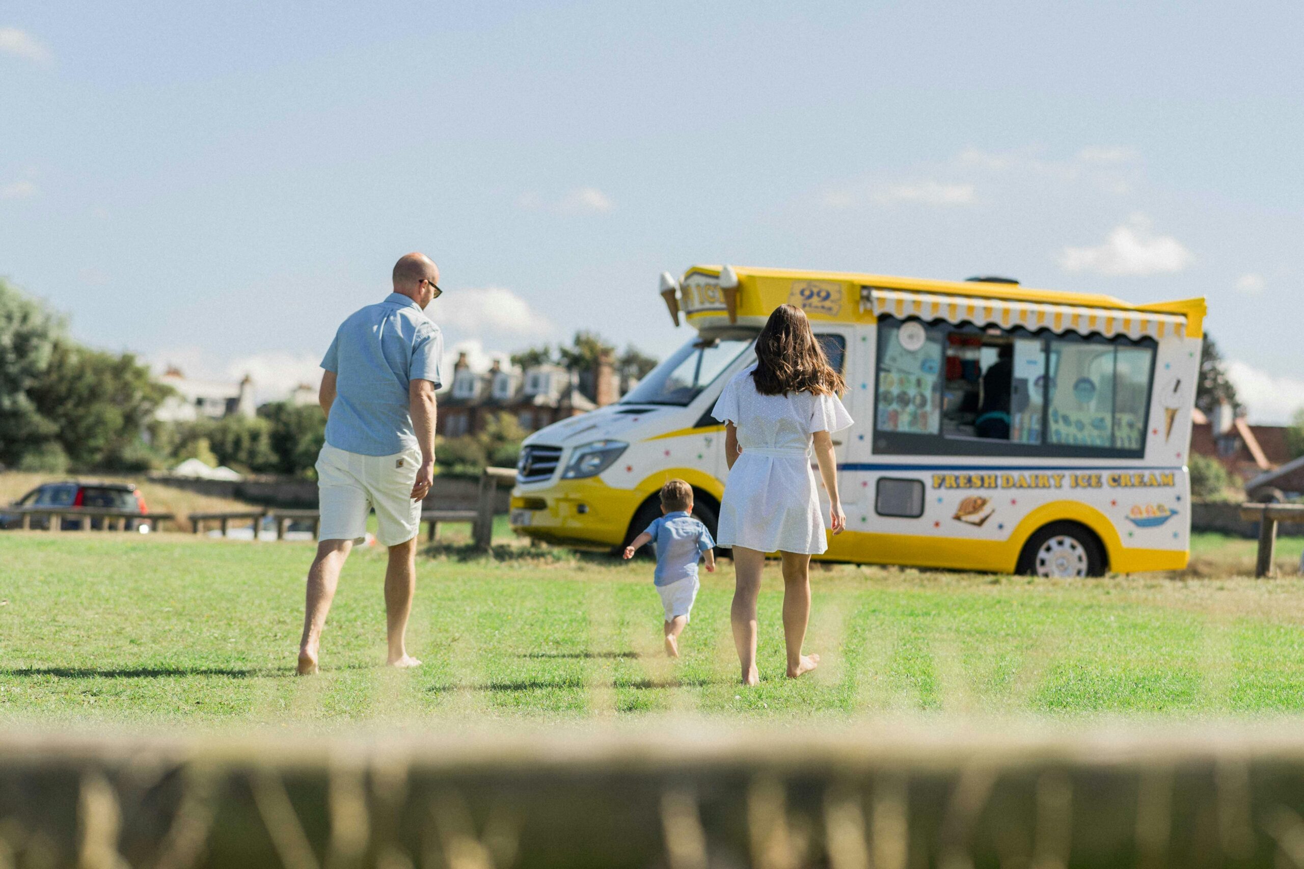 Full image of On a verdant manicured lawn under a summer sky, two parents walk barefoot after their young child, who is running towards a traditionally-coloured yellow-and-white British ice cream van with visible branding reading 'fresh dairy ice cream' and '99 flake'.