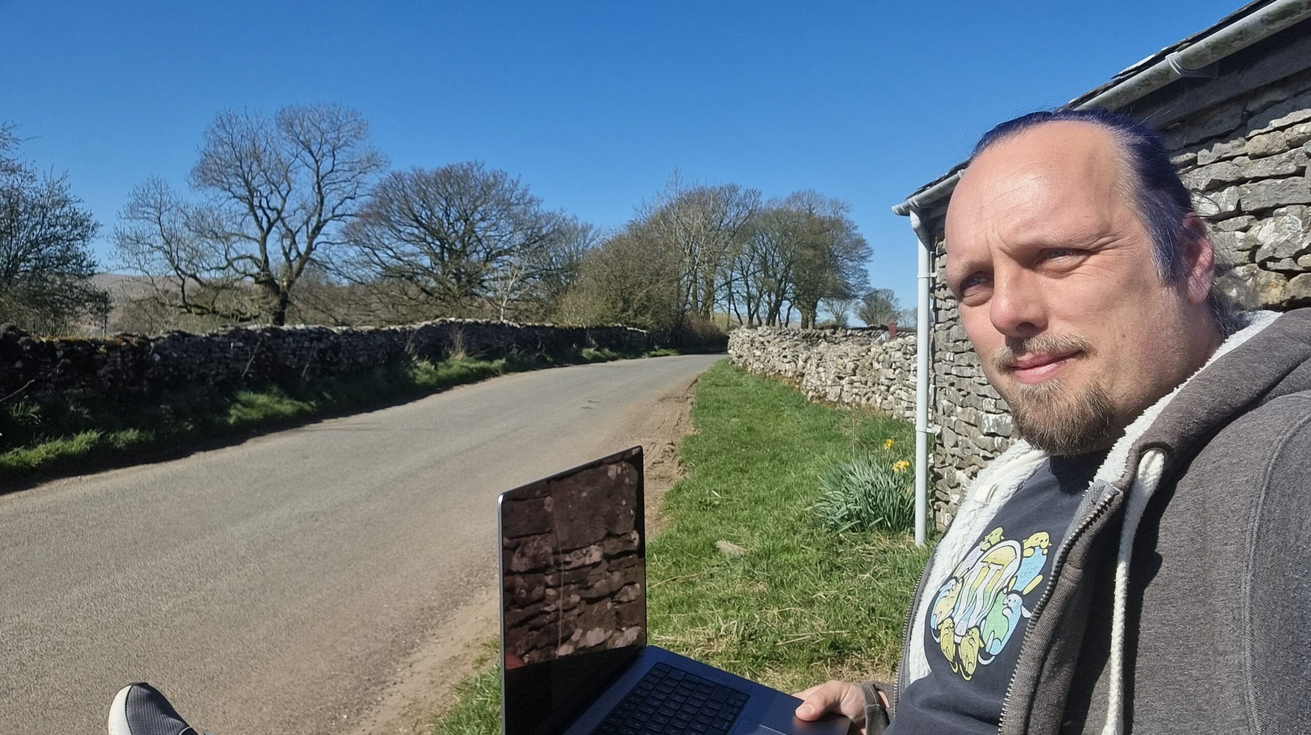 Full image of Dan, with a laptop, sitting alongside a rural Lake District lane in bright sunshine.