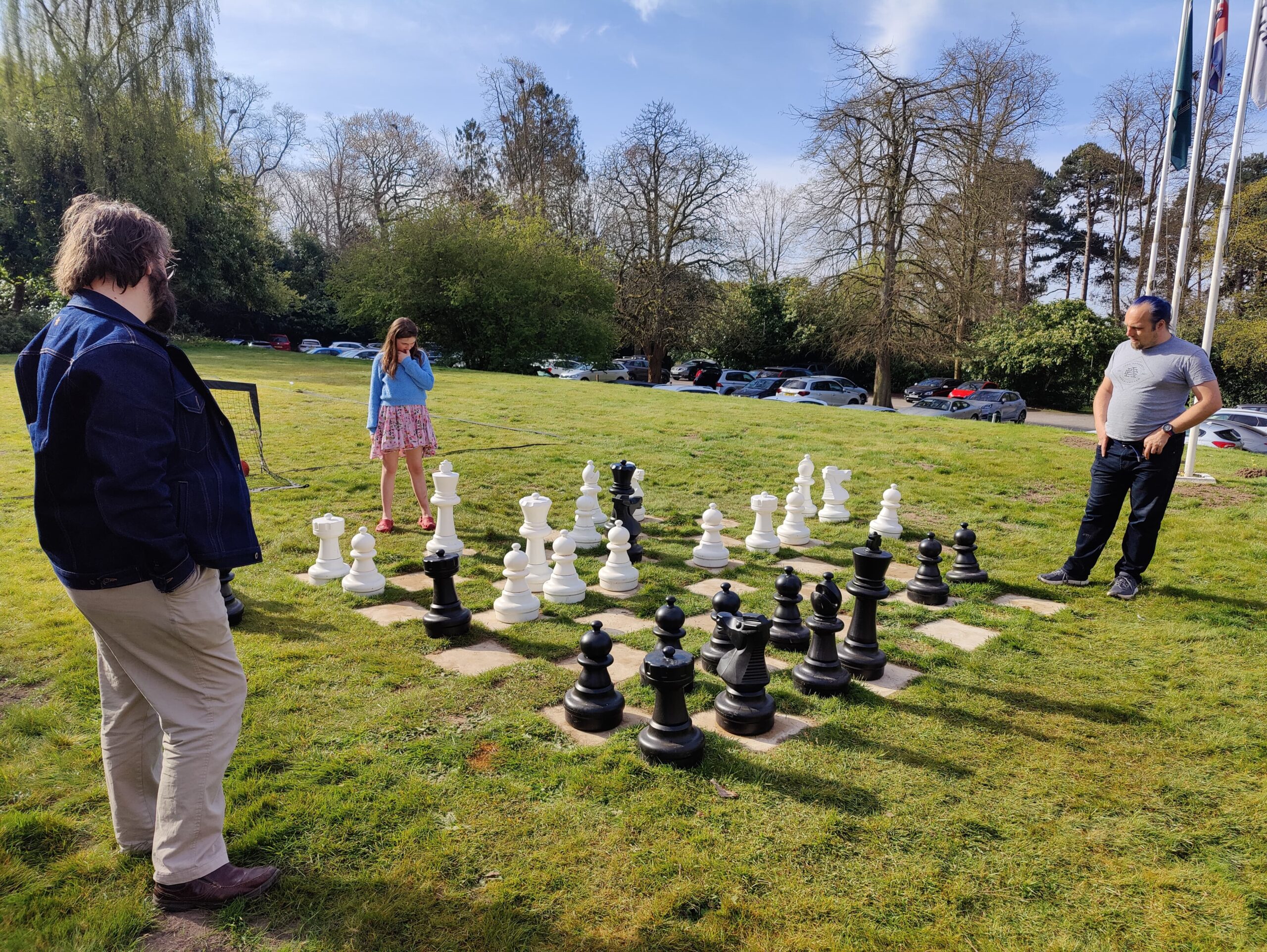 Full image of A girl studies a chessboard on a lawn, watched over by two men: Dan and JTA.