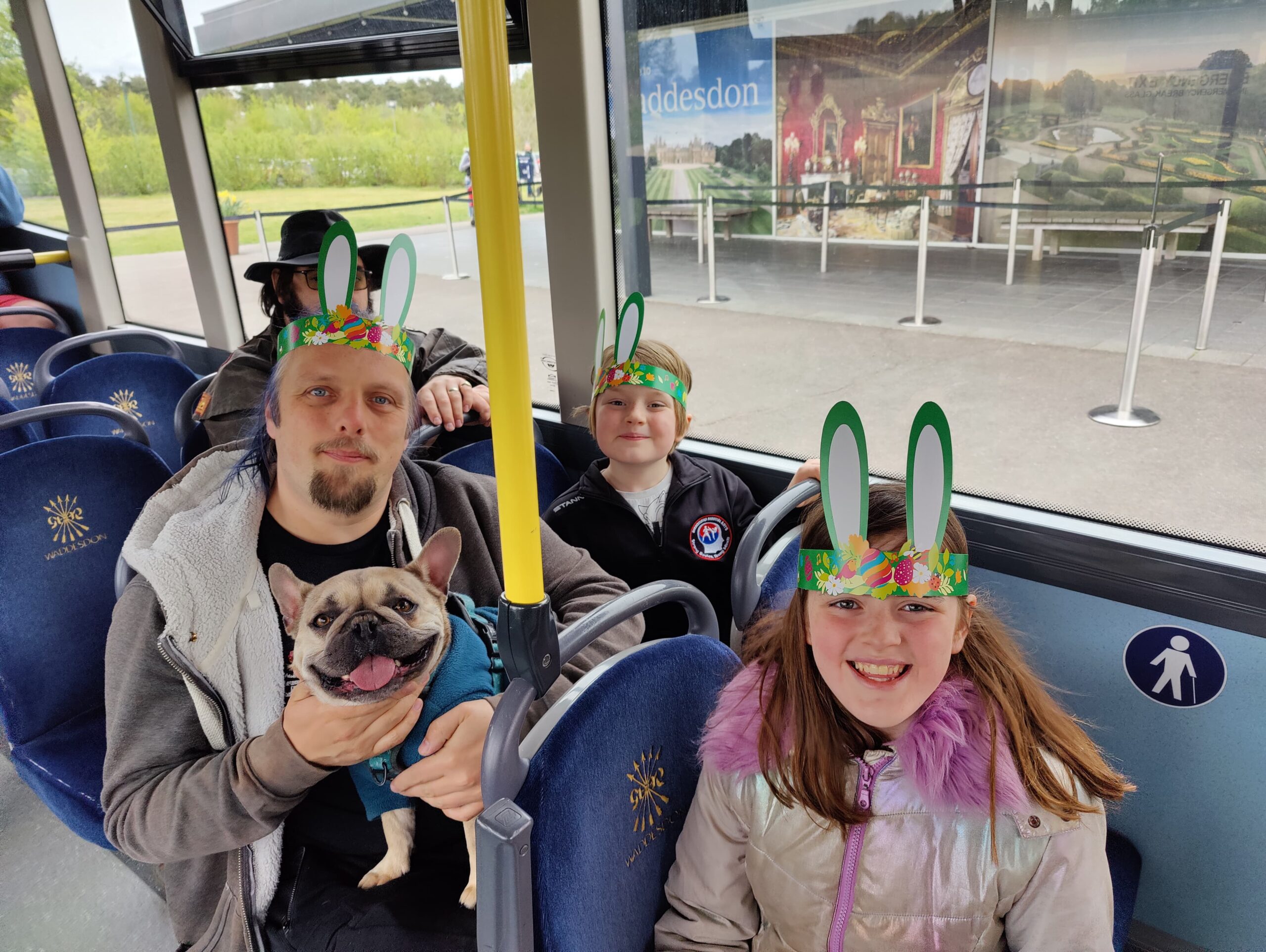 Full image of Dan sits on a bus, a dog in his lap, alongside a boy and a girl. All three humans are wearing cardboard 'rabbit ears' hats.