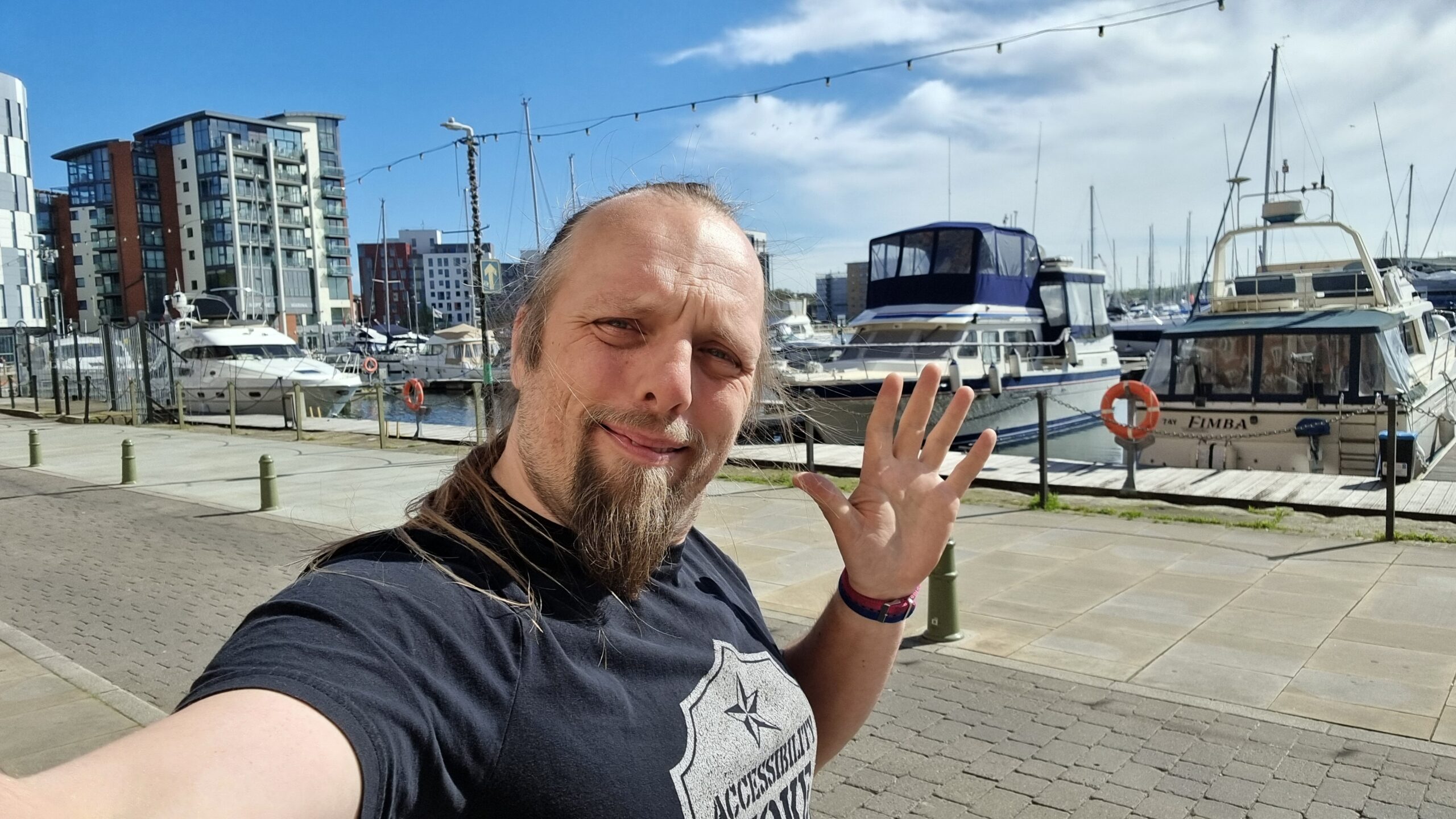 Full image of Dan waves to the camera in front of a pleasure marina and revitalised docklands.