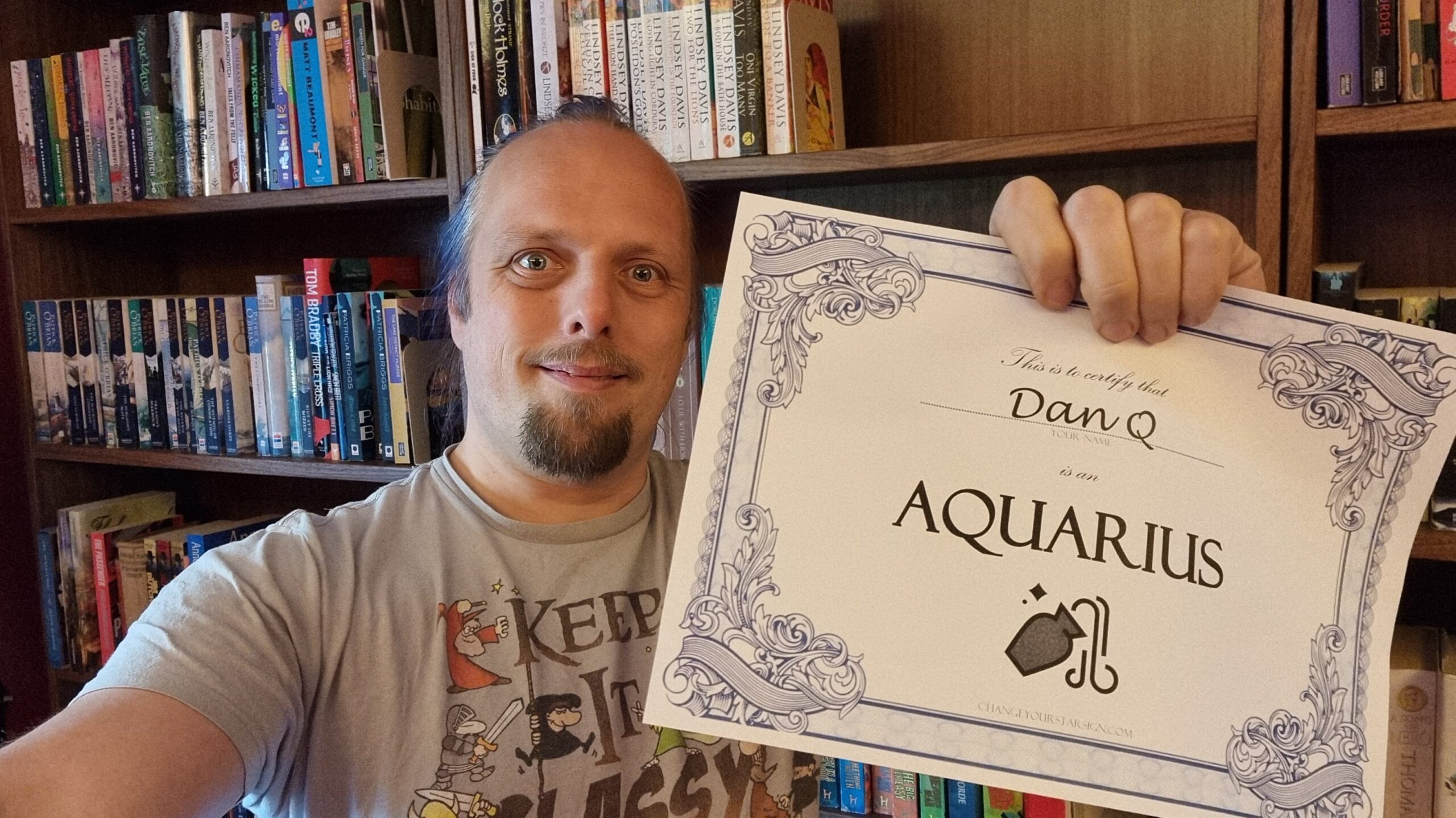 Full image of Dan, in a library, smiles as he holds up a certificate that certifies that he is an Aquarius.