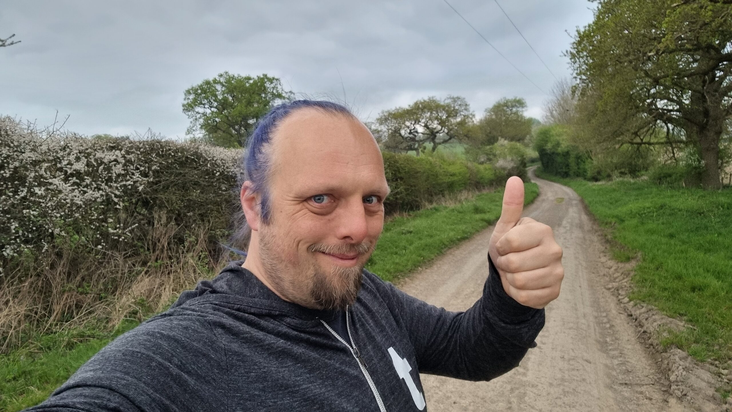 Full image of Dan, with blue hair, throws a thumbs-up for the camera as he stands on a rural countryside lane.