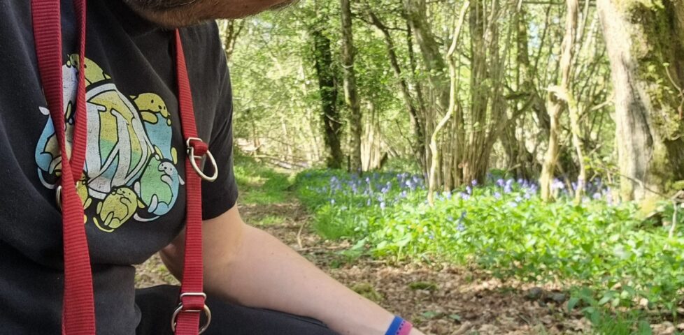 Dan, a dog lead hanging around his neck, crouches alongside his French Bulldog on a trail in a deciduous forest.