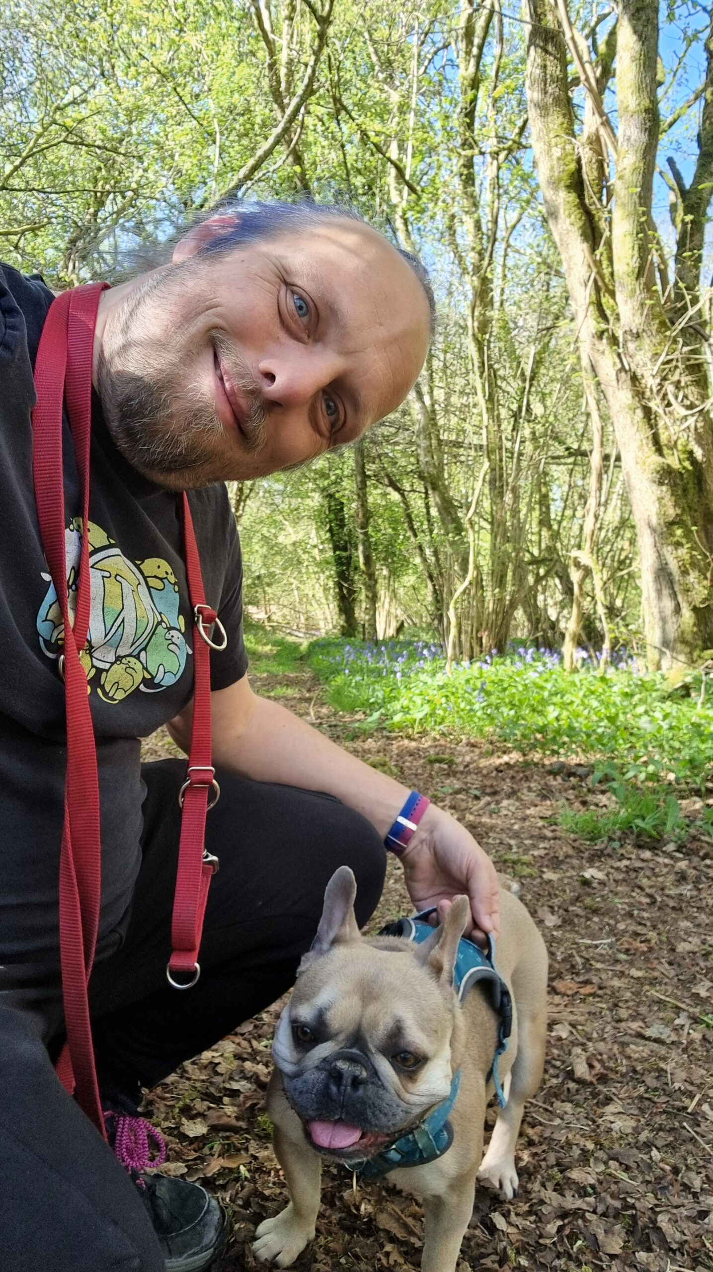 Full image of Dan, a dog lead hanging around his neck, crouches alongside his French Bulldog on a trail in a deciduous forest.