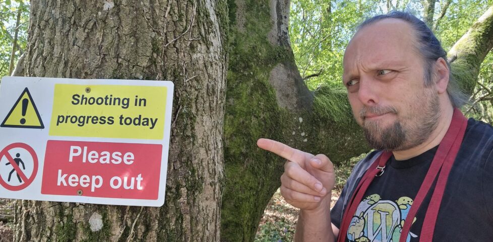 Dan points to a sign screwed to a tree, which reads 'Shooting in progress today' and 'Please keep out'.