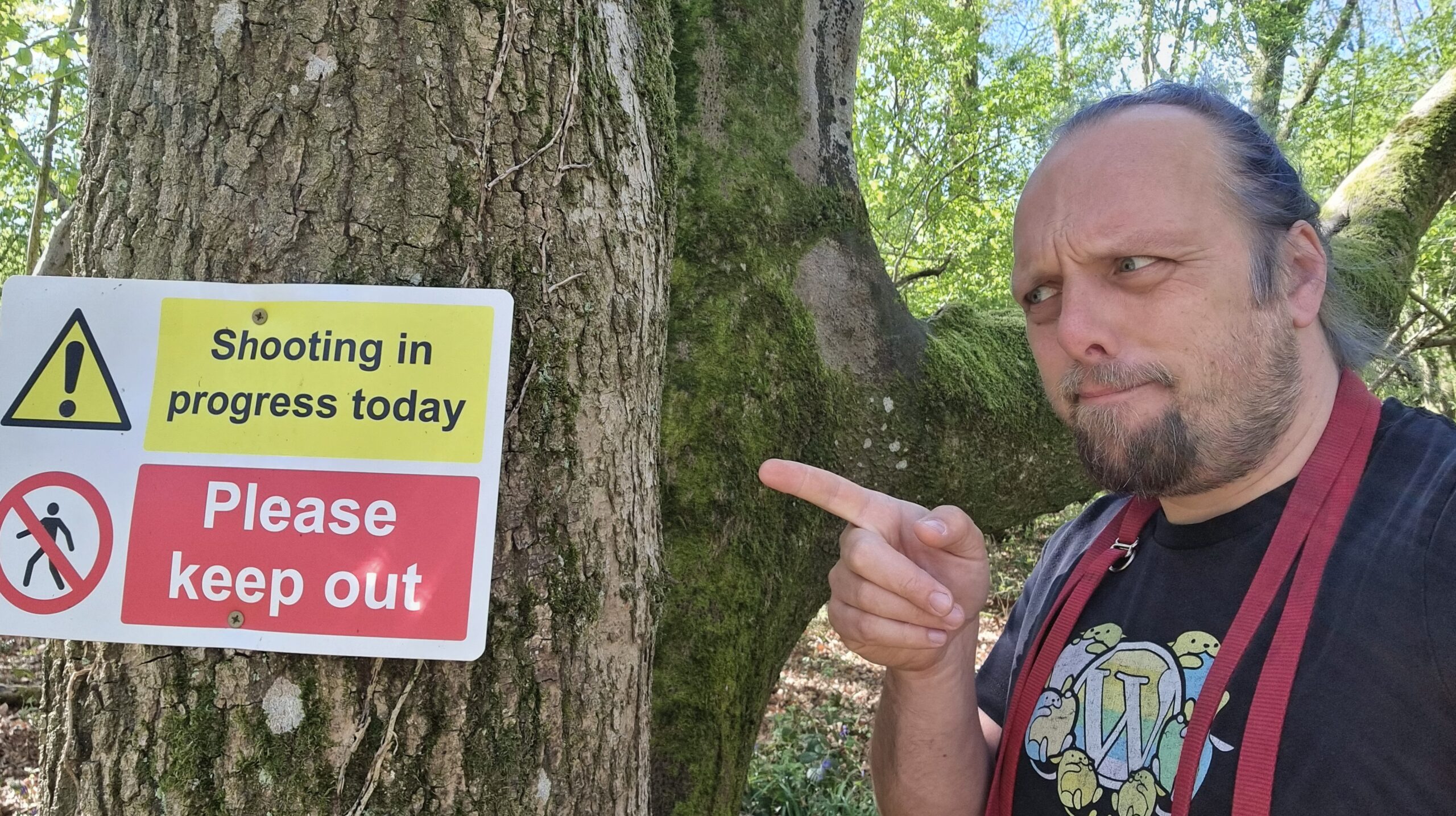 Full image of Dan points to a sign screwed to a tree, which reads 'Shooting in progress today' and 'Please keep out'.