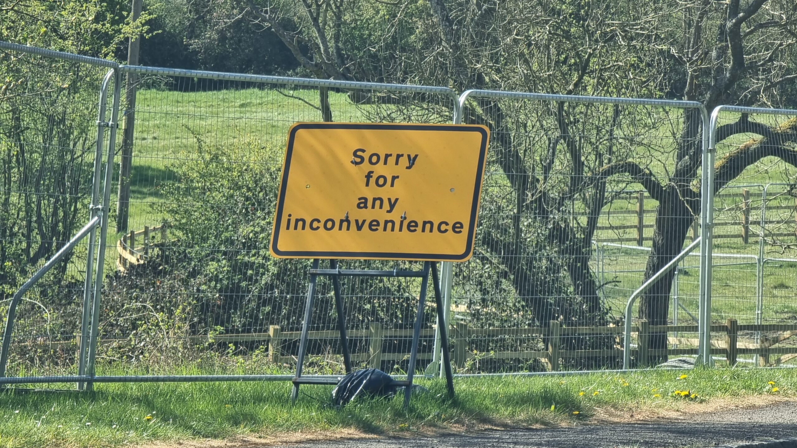 Full image of On a grassy roadside verge, next to a temporary wire fence, a yellow-and-black metal sign reads 'Sorry for any inconvenience'.