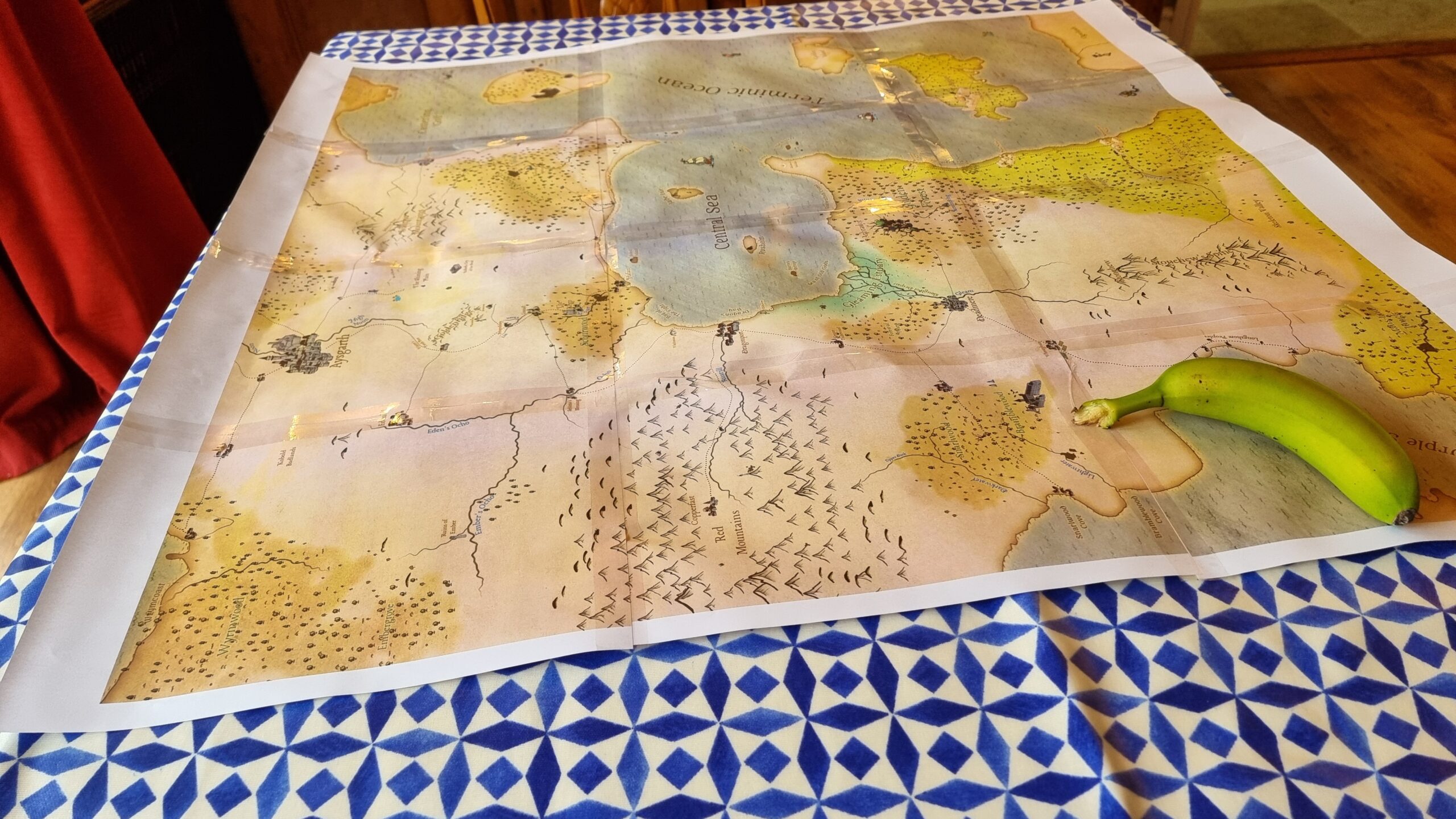 Full image of On a dining table lies a old-style map comprised of 12 sheets of A4 paper, sellotaped together. The map shows the 'Central Sea', an inlet from the 'Terminic Ocean', around which various settlements, forests, mountain ranges, and swamps can be found. An underripe banana sits in one corner of the map, weighing it down.