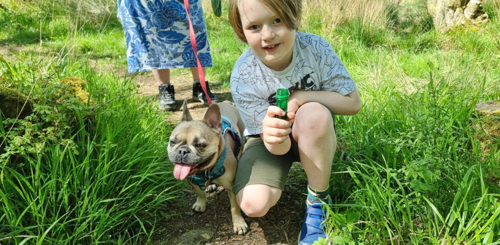 An 8-year-old boy in a white t-shirt and shorts crouches on a grassy trail, alongside a panting French Bulldog. The boy is holding a large green test tube.