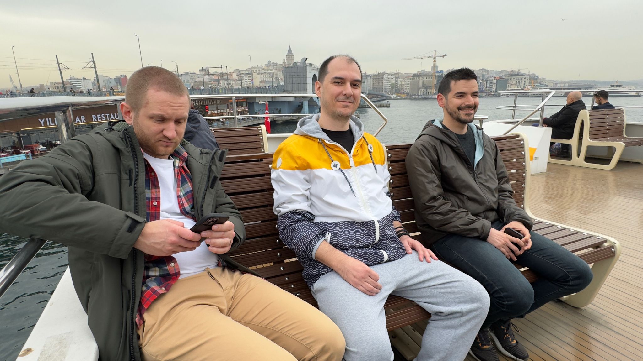 Full image of Three men sit on the upper deck of a passenger ferry near a bridge. A city skyline can be seen in the background.