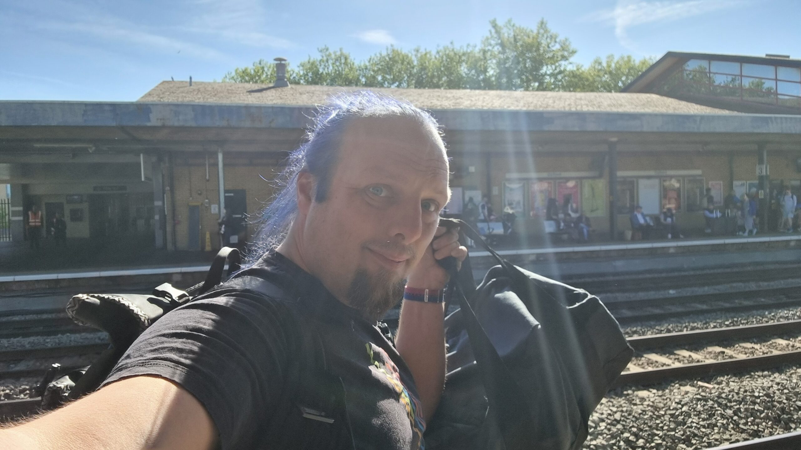 Full image of Dan, with blue hair and wearing a black t-shirt, stands on a sunny train platform holding aloft a medium-sized tote bag.