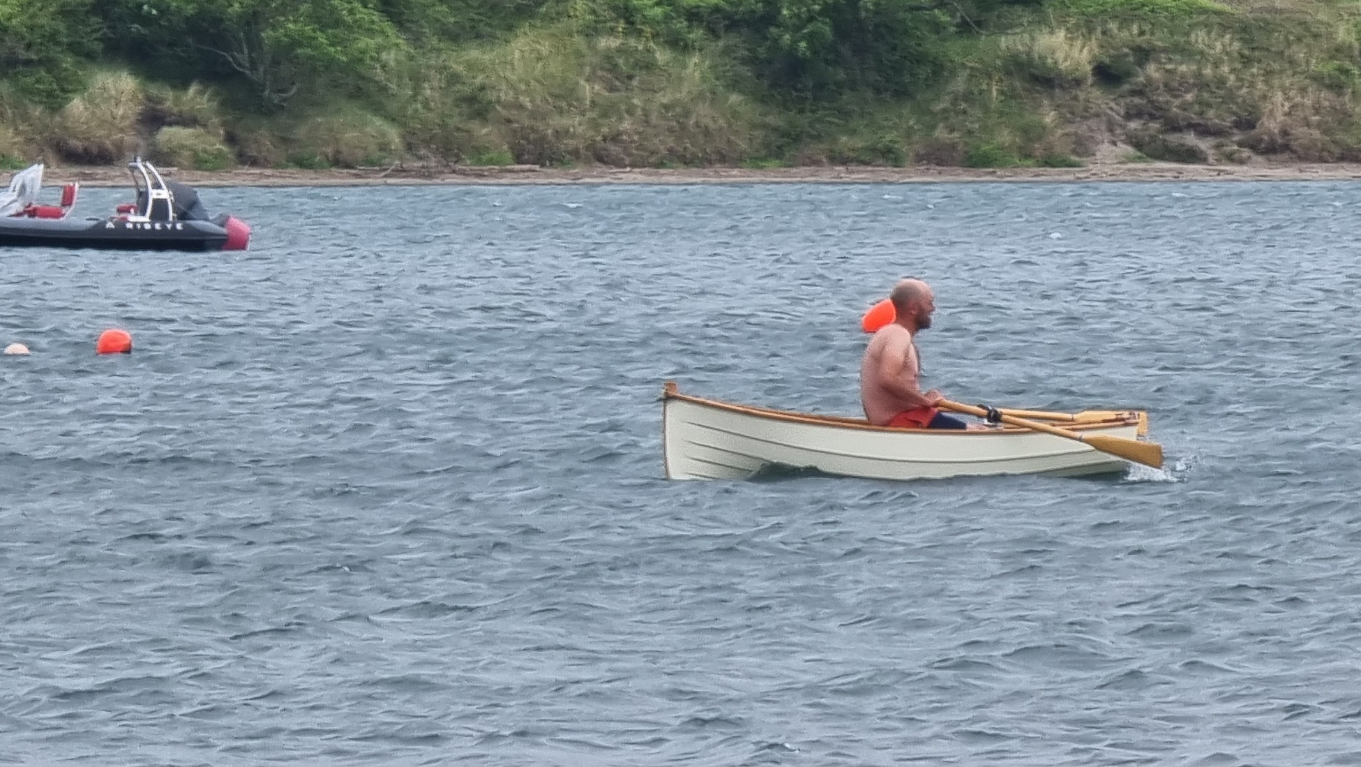 Full image of A shirtless white bald man rows a small white boat past bouys in a choppy estuary.