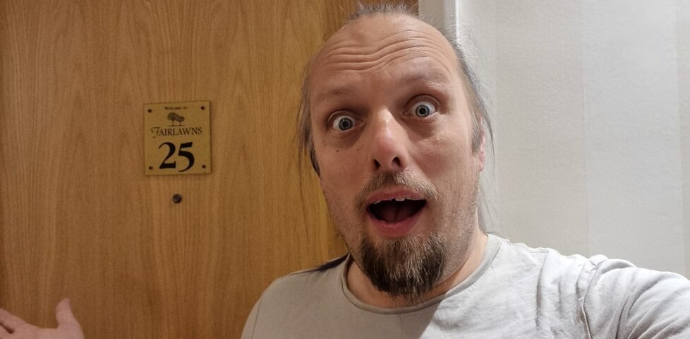 Dan looks shocked as he stands in front of a wooden door on which a brass plaque reads 'Welcome to Fairlawns 25'.