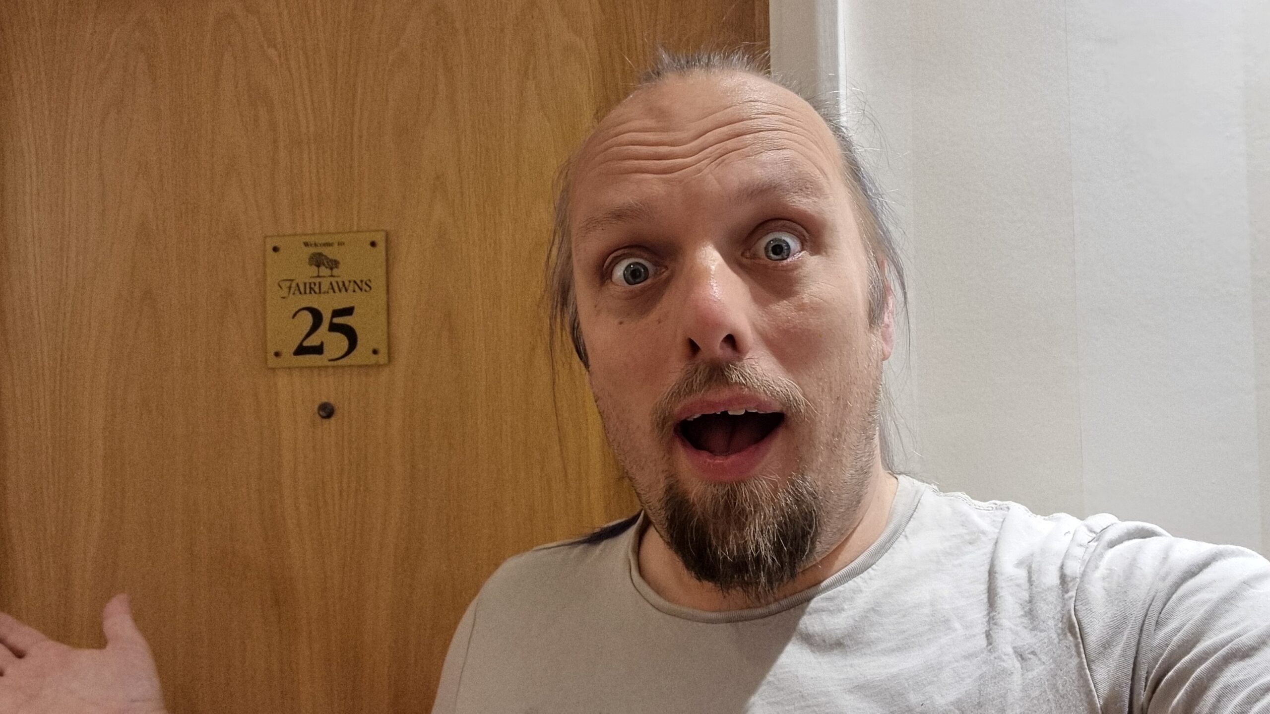 Full image of Dan looks shocked as he stands in front of a wooden door on which a brass plaque reads 'Welcome to Fairlawns 25'.