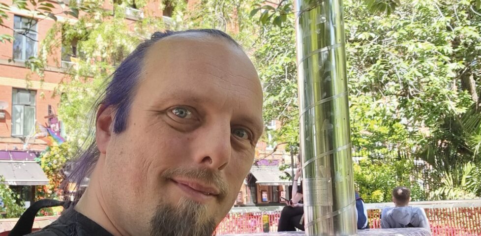 Dan, wearing a pride rainbow t-shirt, stands alongside a steel beacon in the corner of an urban park, under bright late-spring sunlight.