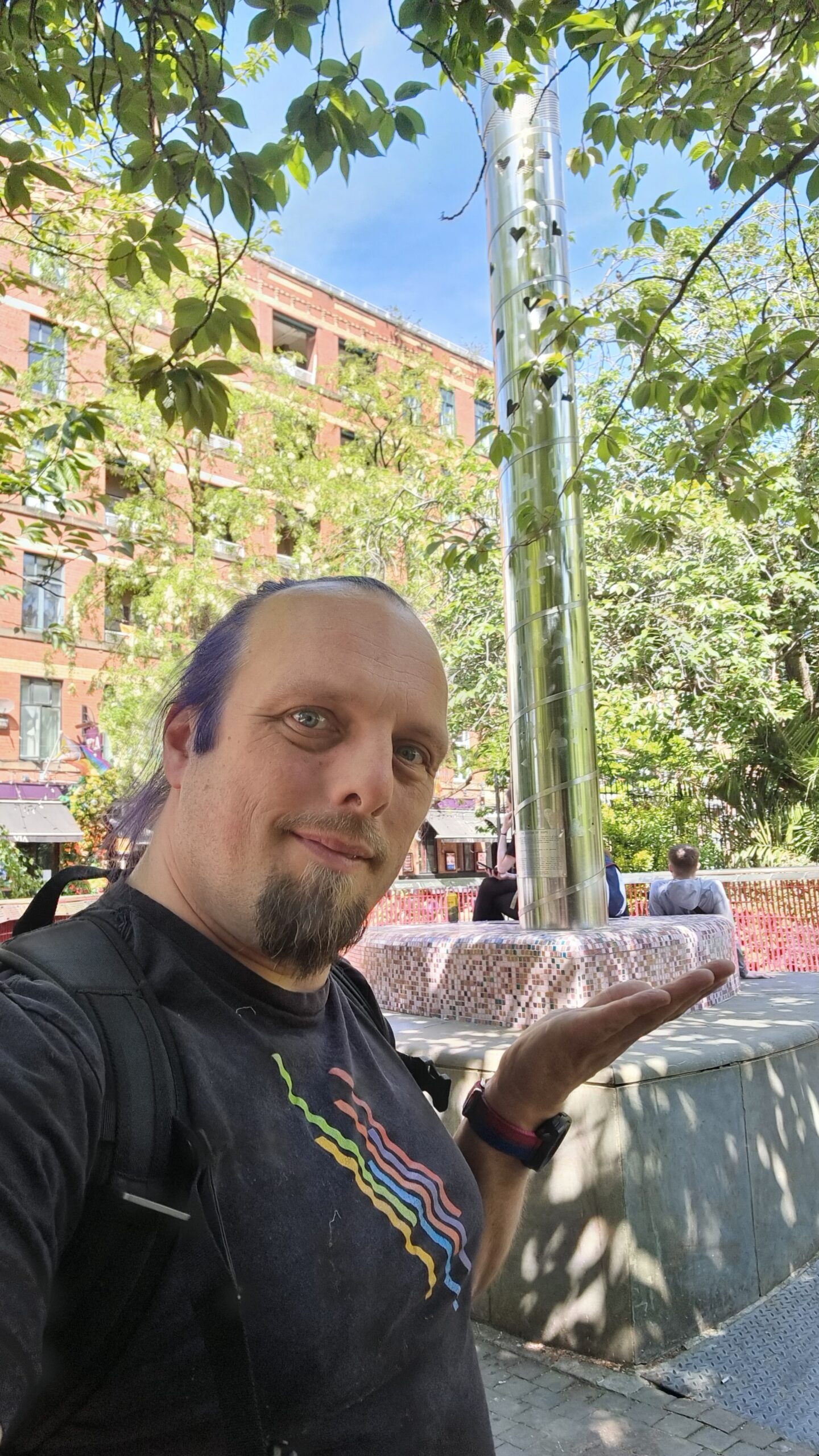 Full image of Dan, wearing a pride rainbow t-shirt, stands alongside a steel beacon in the corner of an urban park, under bright late-spring sunlight.
