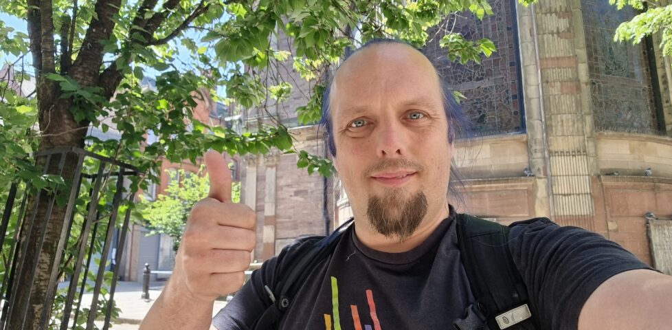 Under a shady tree and in front of a church, Dan throws a thumbs-up.