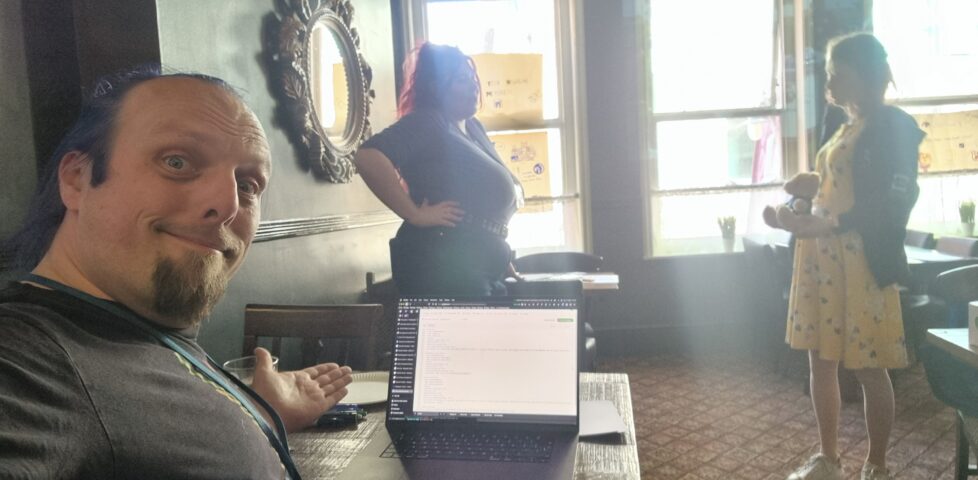 Dan gestures at his laptop in a quiet pub function room, on which he's writing some code. In the background, two women are having a conversation.