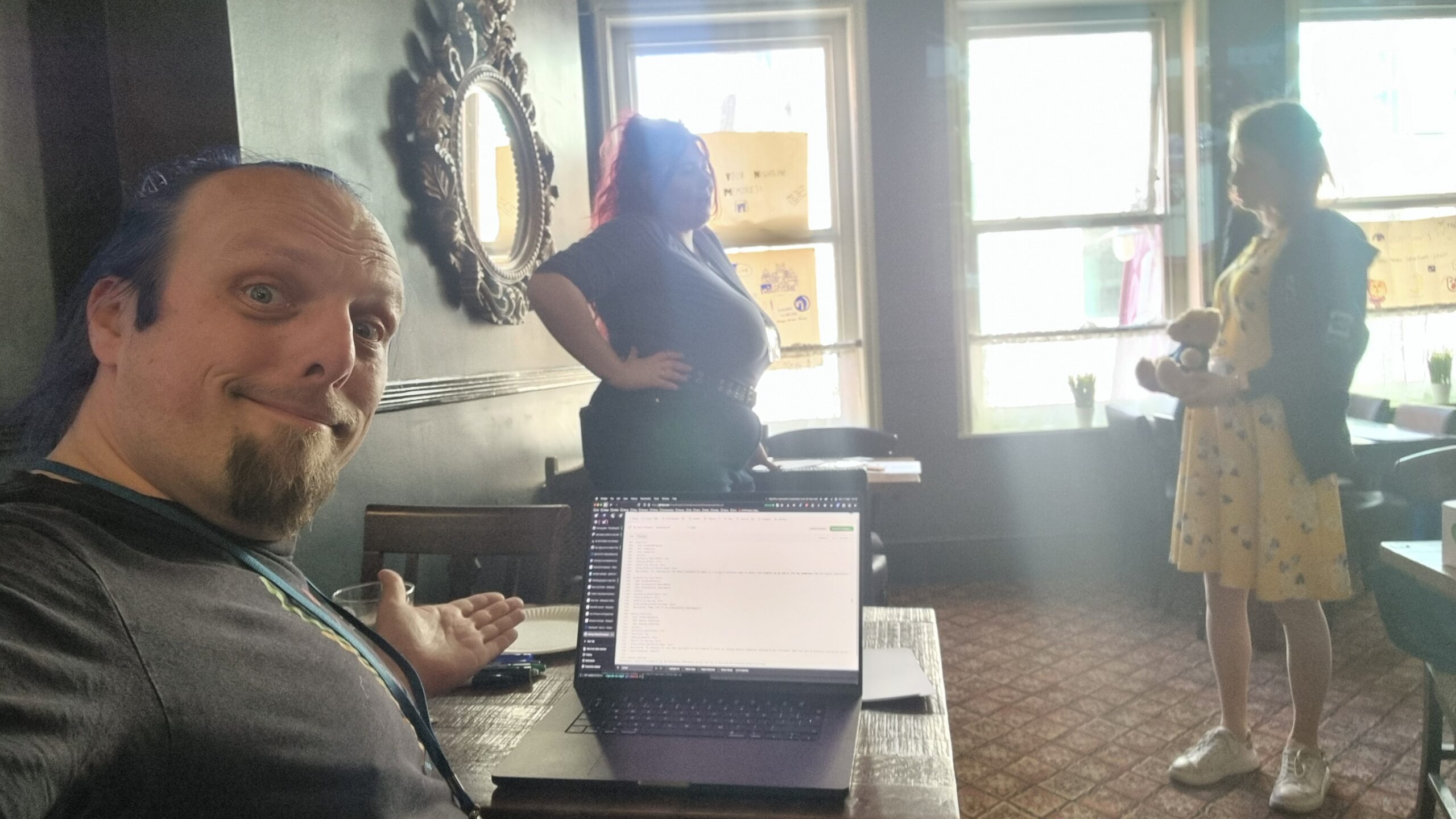 Full image of Dan gestures at his laptop in a quiet pub function room, on which he's writing some code. In the background, two women are having a conversation.