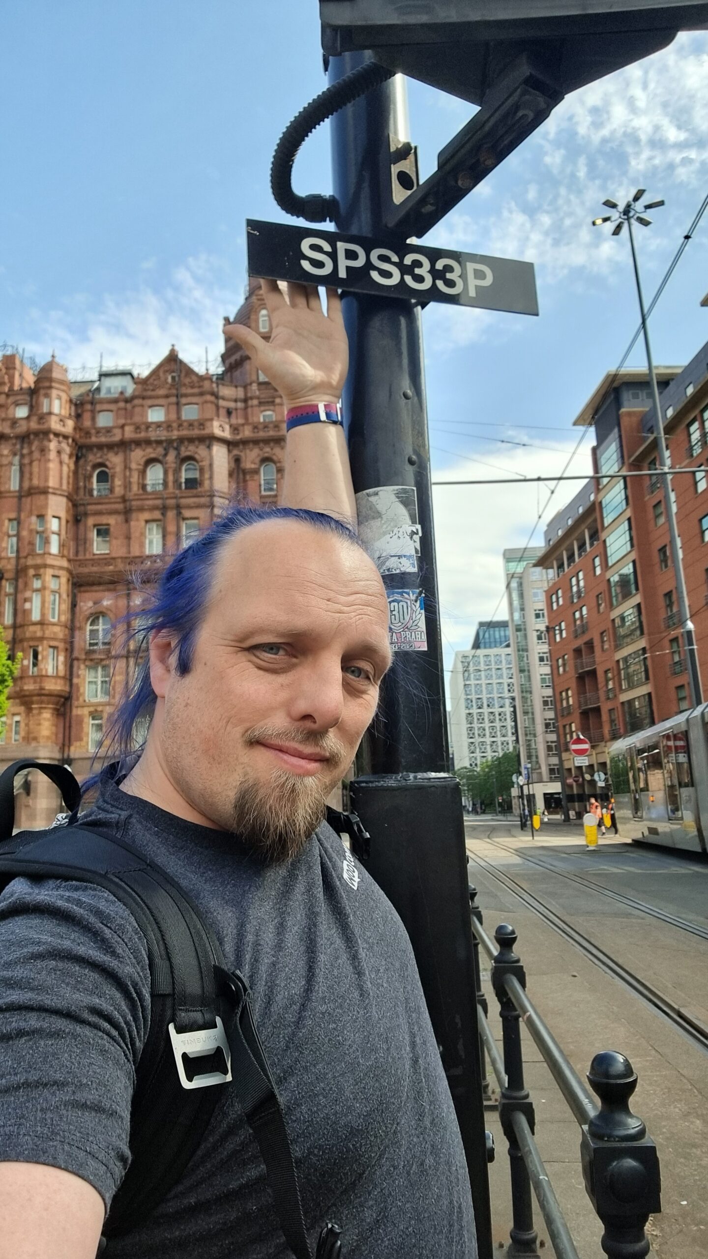 Full image of Dan stretches high to touch the back of a sign mounted on a pole, alongside tram signals, in a city centre.