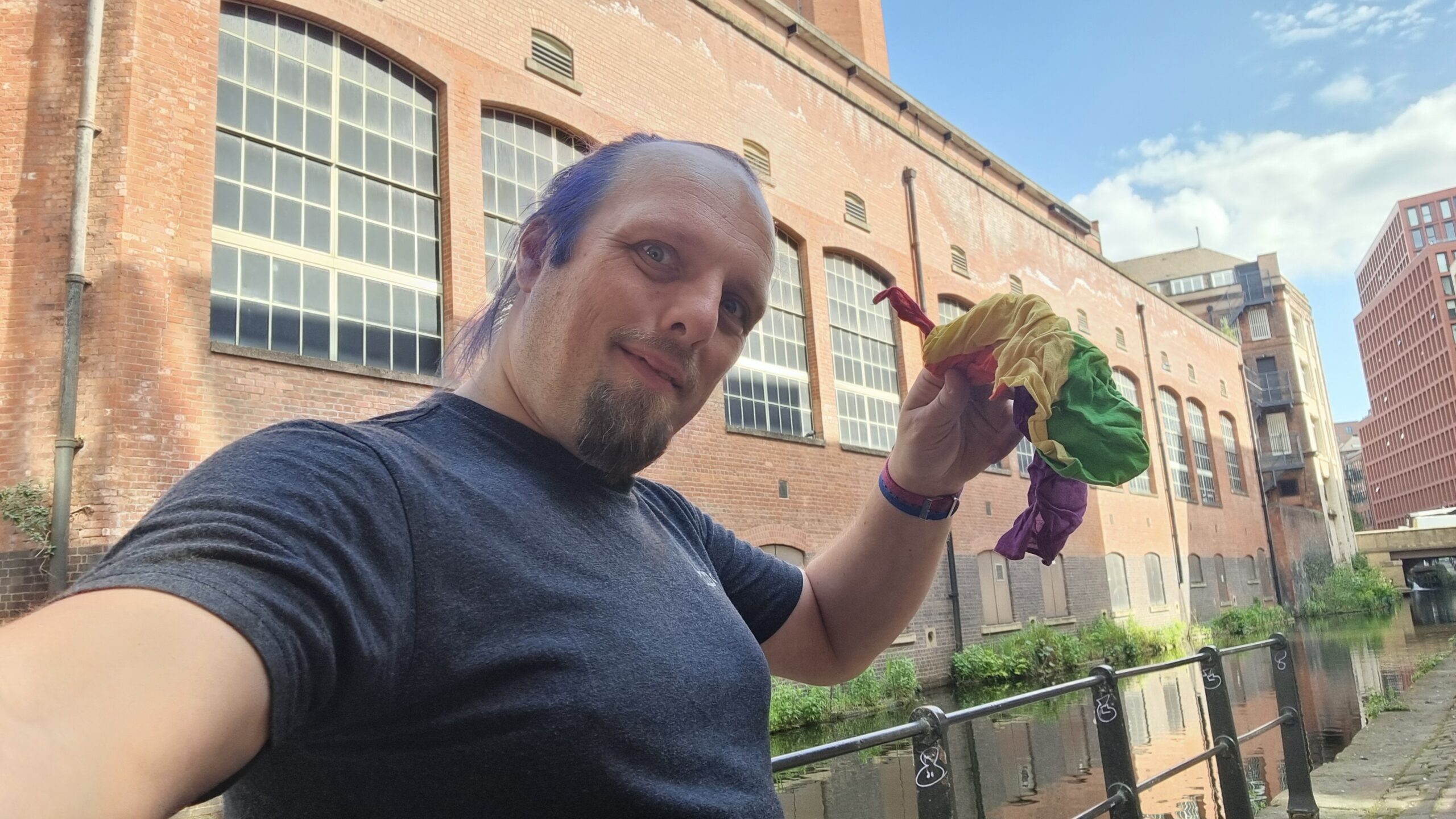 Full image of Dan stands on a reclaimed post-industrial canalside, waving a rainbow-striped bandana.