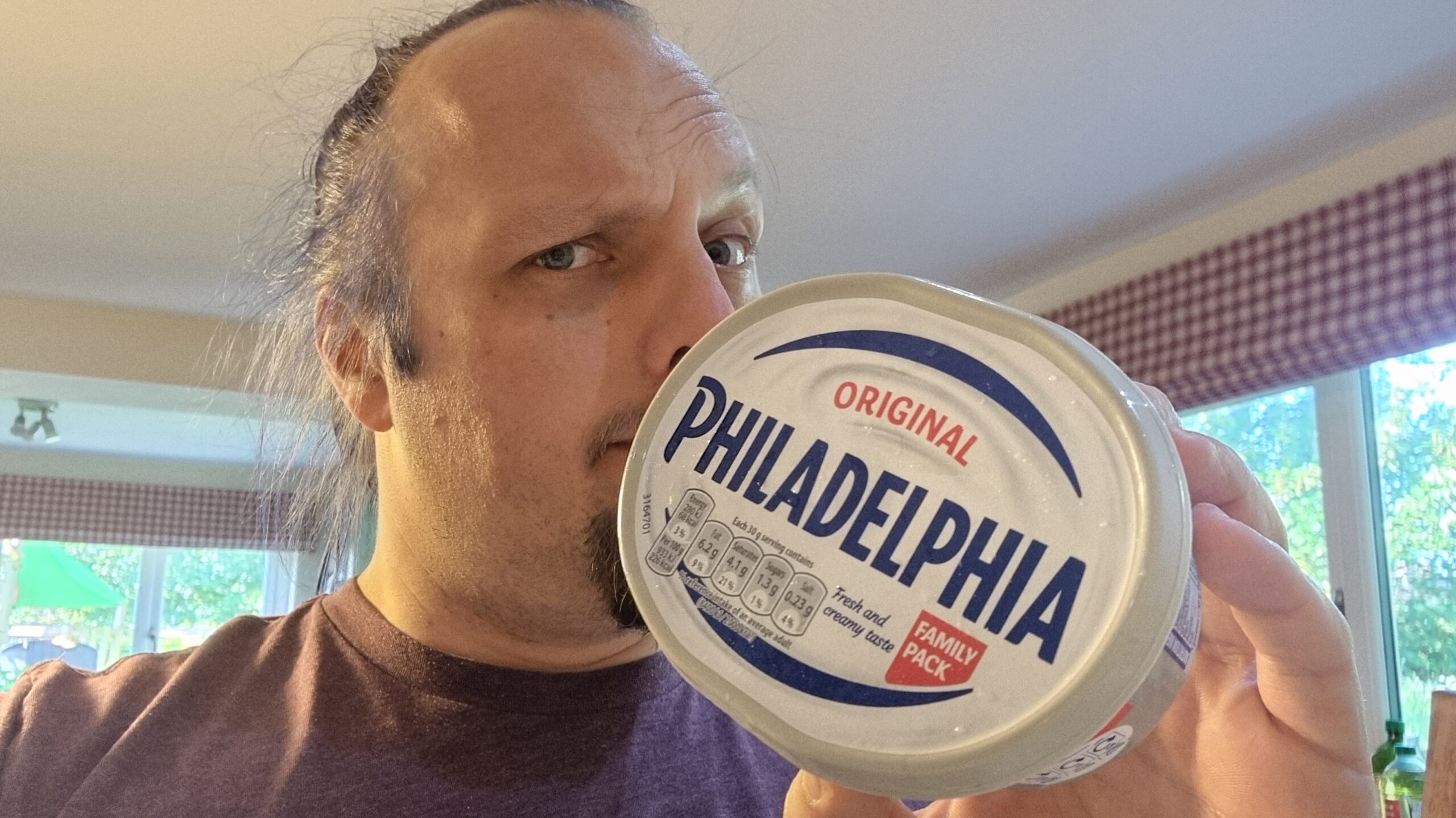Full image of Dan, a white man with blue hair and a goatee, wearing a purple t-shirt, stands in a spacious residential kitchen holding a 'Family Pack'-sized tub of 'Original Philadelphia'.