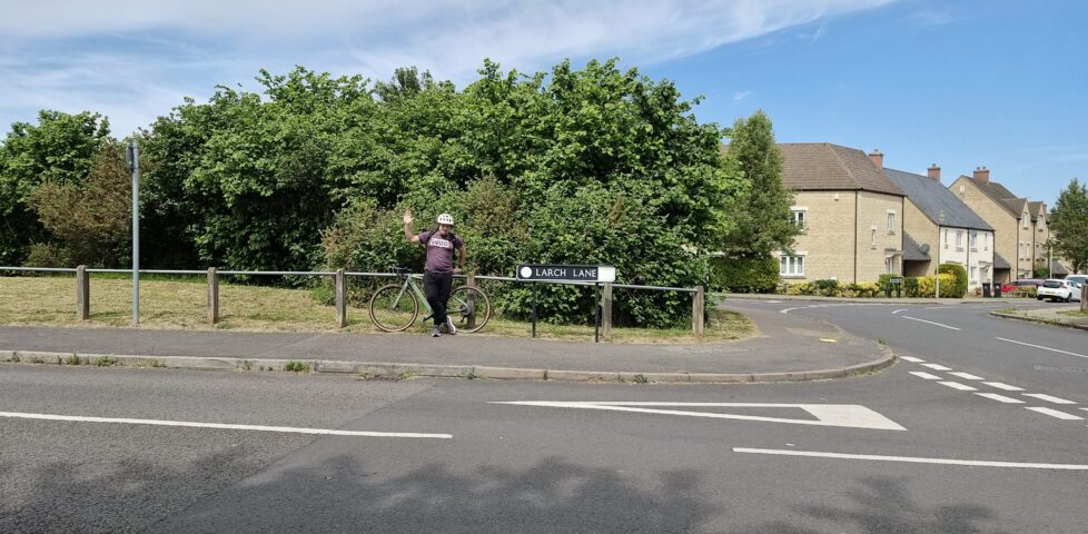 At a suburban junction between two residential streets, near a small green space, Dan waves as he leans against his bicycle.