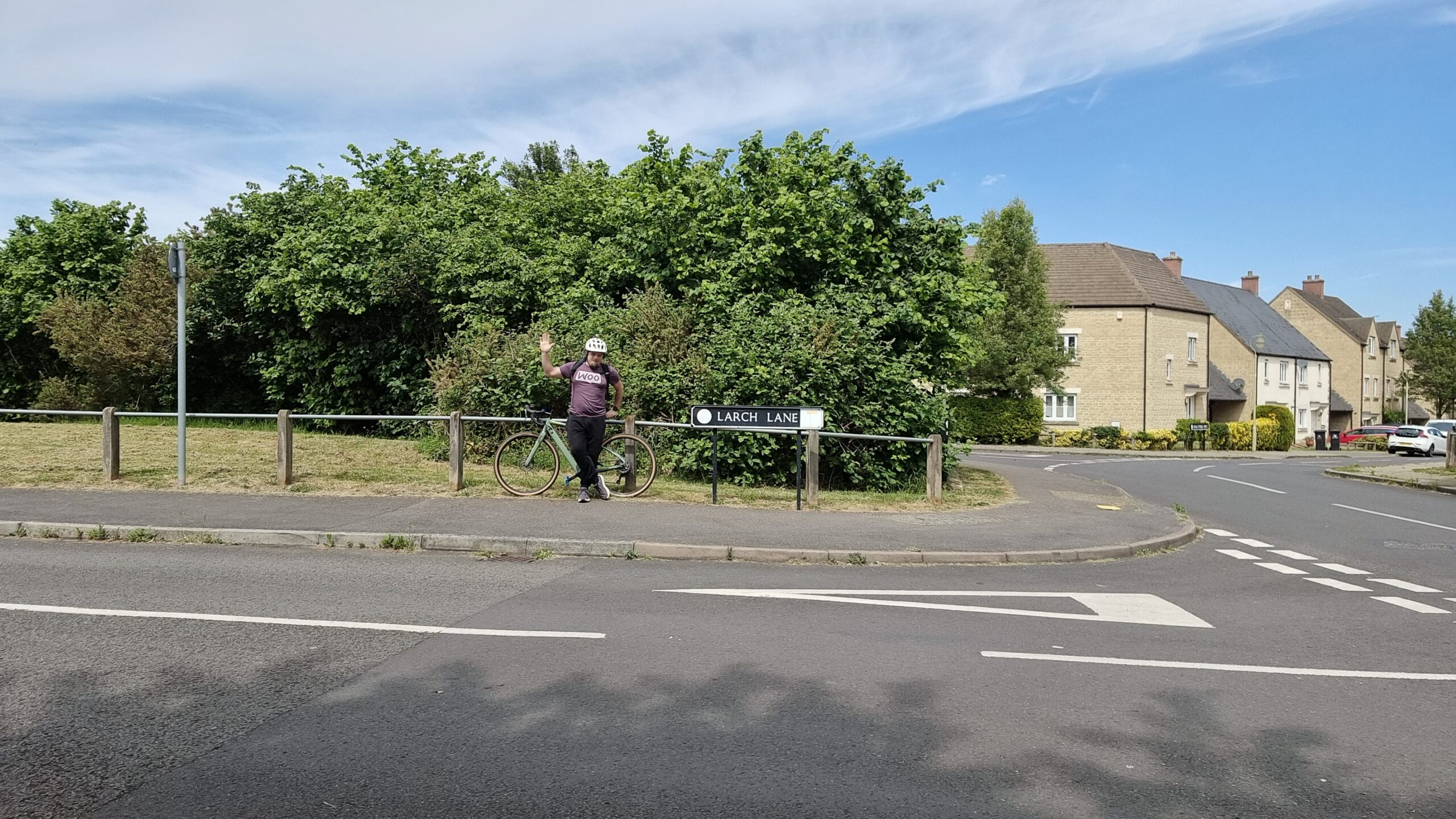Full image of At a suburban junction between two residential streets, near a small green space, Dan waves as he leans against his bicycle.