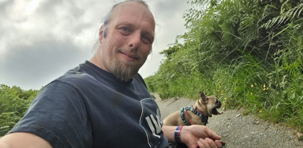 Dan crouches on a path, flanked by tall grass. Behind him, a French Bulldog yawns, her mouth wide and her tongue out.