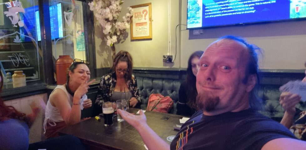 Dan shrugs for the camera as he sits at a pub table with a variety of people; one woman, sat to his left, throws a V-sign with her fingers.