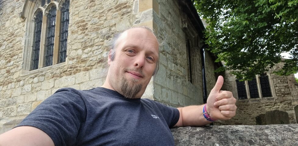 Outside a Norman-style church, Dan throws a thumbs-up sign.