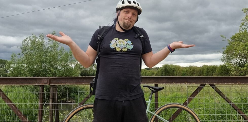 Dan, standing by his bike on an iron footbridge over marshy ground, makes an exaggerated shrugging gesture.