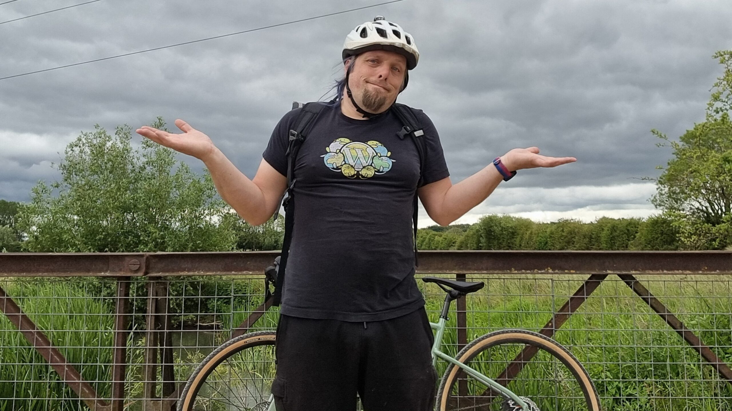Full image of Dan, standing by his bike on an iron footbridge over marshy ground, makes an exaggerated shrugging gesture.
