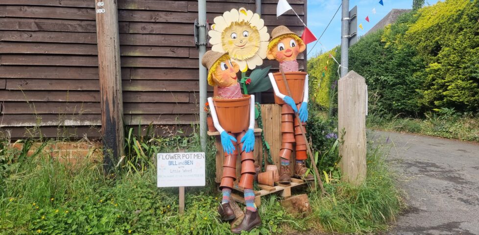 Human-sized scarecrows of classic childrens' TV characters Bill and Ben the Flower Pot Men and their friend Little Weed, constructed mostly out of plant pots, standing at the corner of a road by a wood-panelled outbuilding.
