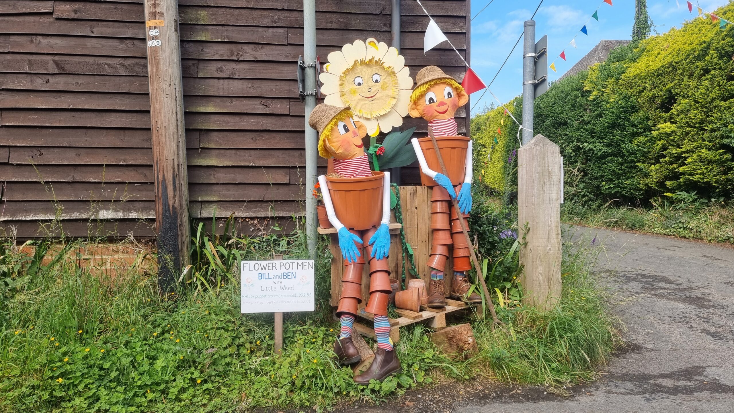 Full image of Human-sized scarecrows of classic childrens' TV characters Bill and Ben the Flower Pot Men and their friend Little Weed, constructed mostly out of plant pots, standing at the corner of a road by a wood-panelled outbuilding.