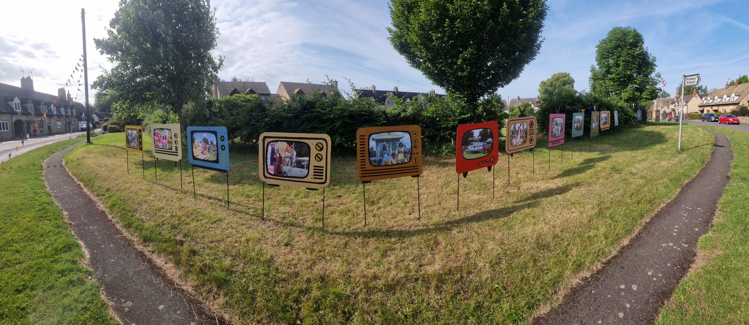 Full image of Panoramic view of a village green, flanked by houses, around which several decorative boards have been erected: made to look like old-fashioned televisions, each shows a photograph of an event in the village's past.