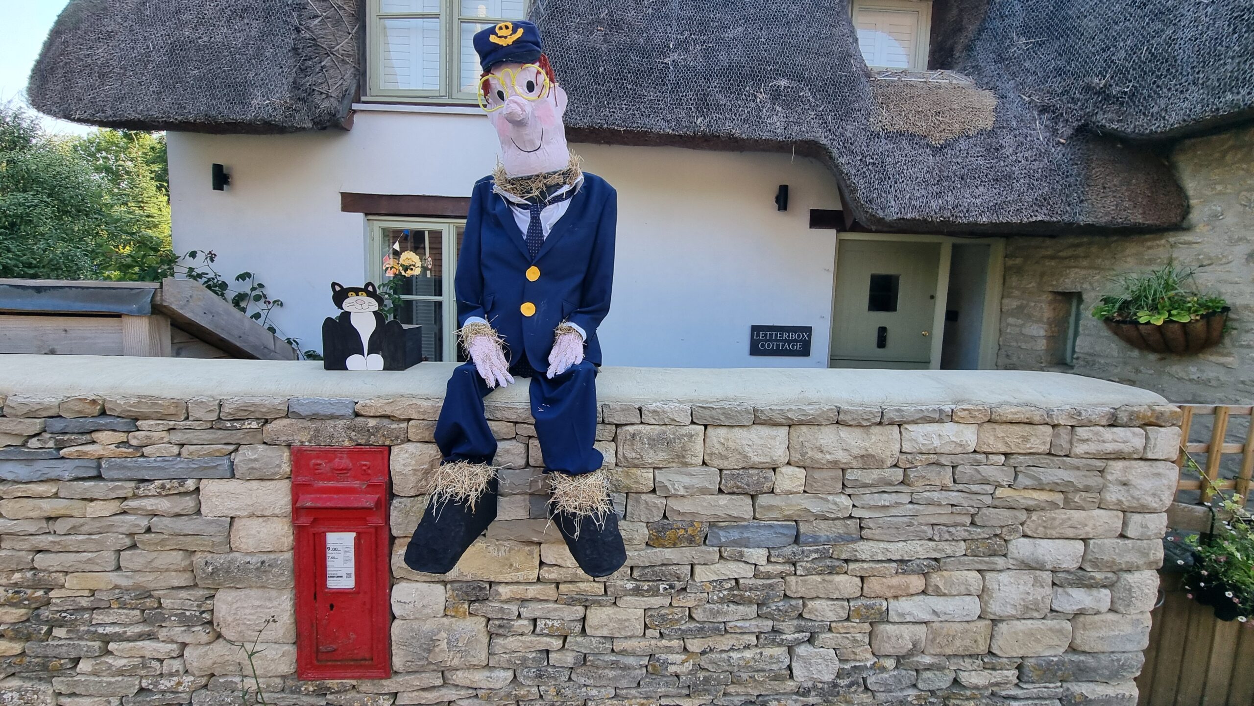 Full image of A scarecrow of Postman Man, alongside a cardboard cutout of Jess (his black-and-white cat), sits atop a stone wall. The wall contains an in-wall Royal Mail postbox, and the thatched house behind is called Letterbox Cottage, contributing to the theme for this scarecrow.