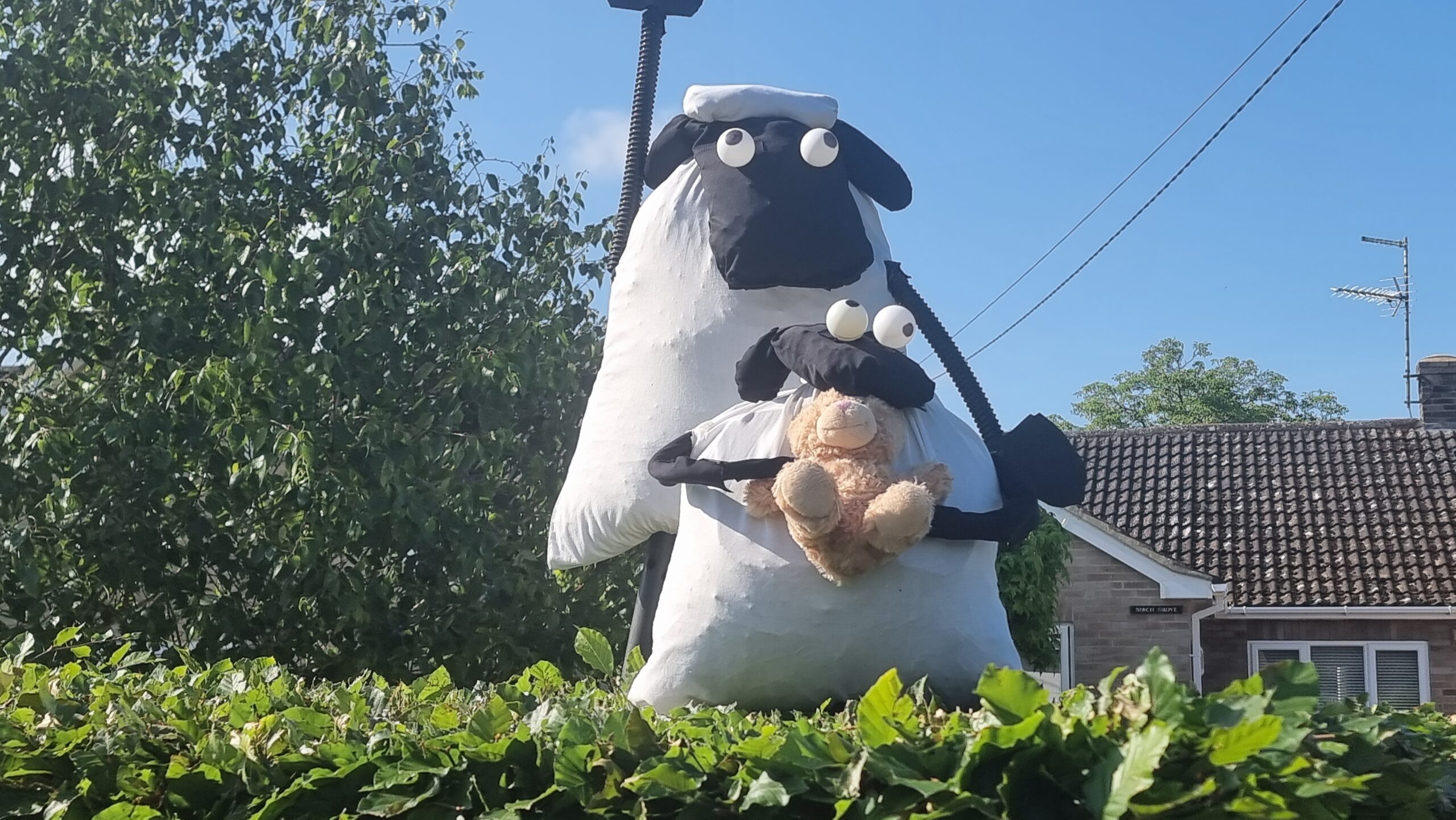 Full image of Large scarecrows of two anthropomorphic sheep, the smaller one holding a teddy bear, stood atop a hedgerow.